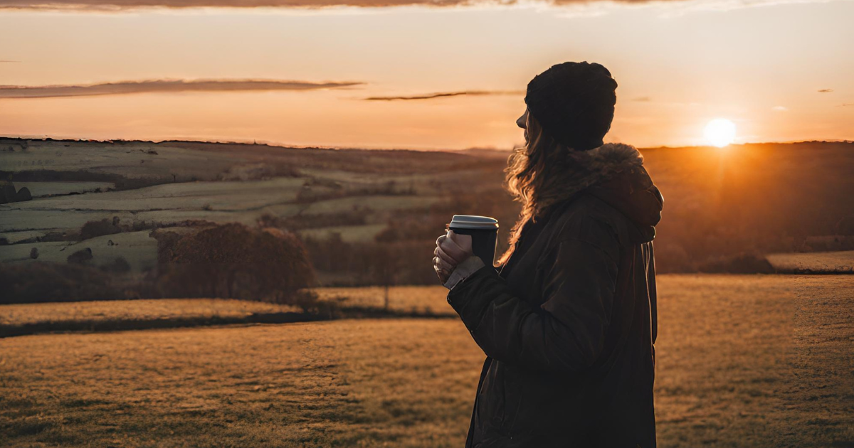 woman holding a coffee cup looking at the sun set over the hills